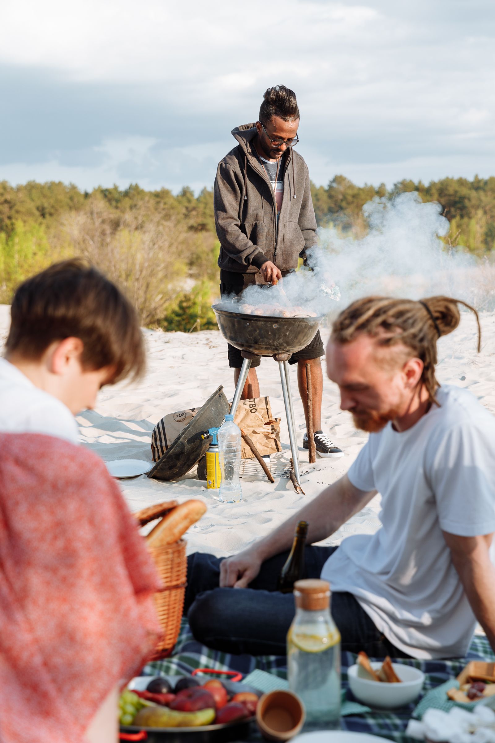 Friends gathered on a beach picnic blanket while one person grills nearby